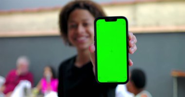 Kameraya yakın yeşil ekran krom anahtarı olan akıllı telefonu tutan, avlu ortamında gülümseyen, arka planda bulanık insanlar olan genç bir çocuk.
