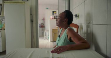 Yaşlı siyah bir kadın, rahat bir mutfakta yemek masasında oturmuş, düşünceli bir şekilde uzaktan bakıyor, sıcak ve tanıdık bir evde huzurlu bir anı yakalıyor.