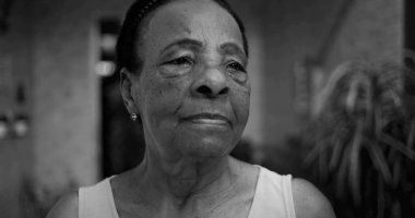 Düşünceli bir profilde yaşlı bir Afro kökenli kadın, kameradan başka tarafa bakıyor, açık bir ev ortamında düşünceli bir yüz ifadesi, doğal ve samimi.