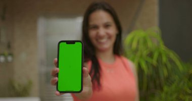Elinde yeşil ekran ve krom tuşlu akıllı telefon tutan kadın kameraya bakıyor, canlı arka plan, dijital veya reklam amaçlı kişiselleştirilebilir aygıt sunuyor