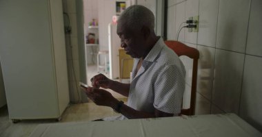 Bir yaşlı siyah adam, sade bir mutfakta yemek masasında oturuyor, akıllı telefon kullanıyor, günlük hayatta sıcak ve tanıdık bir ortamda teknolojiyi ön plana çıkarıyor.