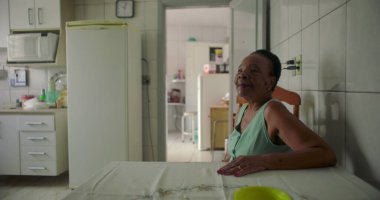 Yaşlı siyah bir kadın, rahat bir mutfakta yemek masasında oturmuş, düşünceli bir şekilde uzaktan bakıyor, sıcak ve tanıdık bir evde huzurlu bir anı yakalıyor.
