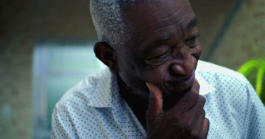 Düşünceli ve biraz endişeli bir ifadeyle yaşlı bir Afro kökenli adam, elleri çenesinde dinleniyor, sakin bir ev ortamında derin düşüncelere dalmış.