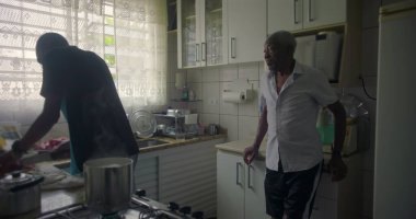 Afrika kökenli bir aile mutfakta yemek hazırlıyor, kadın ocağın yanında, yaşlı bir adam ayakta ve gözlem yapıyor, aile dinamiklerini ve günlük rutinlerini yakalıyor.