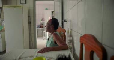 Yaşlı siyah bir kadın, rahat bir mutfakta yemek masasında oturmuş, düşünceli bir şekilde uzaktan bakıyor, sıcak ve tanıdık bir evde huzurlu bir anı yakalıyor.