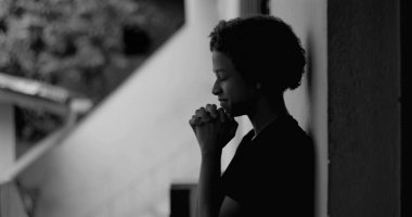 Duvara yaslanmış genç Afrikalı Amerikalı çocuk, dua ederken ellerini sımsıkı tutuyor, rehberlik ve umut arıyor, inanç, esneklik ve monokromdaki iç gücü yansıtıyor.