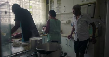 Afrika kökenli bir aile mutfakta yemek hazırlıyor, kadın ocağın yanında, yaşlı bir adam ayakta ve gözlem yapıyor, aile dinamiklerini ve günlük rutinlerini yakalıyor.