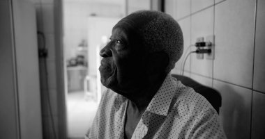 Siyah-beyaz görünümlü yaşlı bir Afro-torunu, düşünceli ve sakin bir ifadeyle yukarı bakıyor, sessiz bir ev ortamında oturuyor