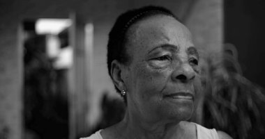 Düşünceli bir profilde yaşlı bir Afro kökenli kadın, kameradan başka tarafa bakıyor, açık bir ev ortamında düşünceli bir yüz ifadesi, doğal ve samimi.