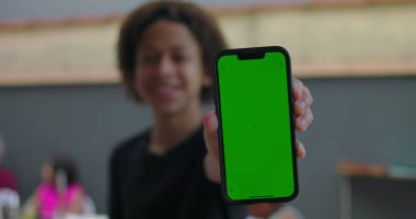 Afrika kökenli bir gencin elinde yeşil ekranlı akıllı telefon, krom anahtar ekranlı, özgüvenle gülümseyen, arka planda çeşitli yetişkinlerle gelişigüzel toplanan