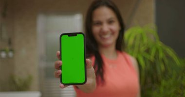 Elinde yeşil ekran ve krom tuşlu akıllı telefon tutan kadın kameraya bakıyor, canlı arka plan, dijital veya reklam amaçlı kişiselleştirilebilir aygıt sunuyor