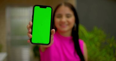 Kameraya yakın yeşil ekran krom anahtarı olan akıllı telefonu tutan genç bir kadın, güvenli bir şekilde gülümsüyor, açık bir evde kişiselleştirilebilir içerik için boş ekranı gösteriyor