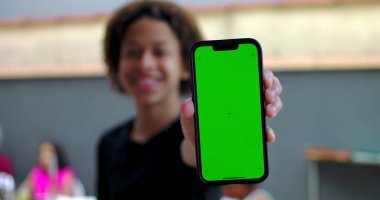 Kameraya yakın yeşil ekran krom anahtarı olan akıllı telefonu tutan, avlu ortamında gülümseyen, arka planda bulanık insanlar olan genç bir çocuk.