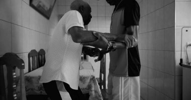 Genç bir adamın yardımıyla yürüyen yaşlı bir adam, ilgi ve destek anı, nesiller arasındaki empati ve bağlantıyı yansıtıyor, siyah ve beyaz.