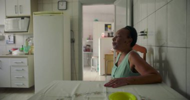 Yaşlı siyah bir kadın, rahat bir mutfakta yemek masasında oturmuş, düşünceli bir şekilde uzaktan bakıyor, sıcak ve tanıdık bir evde huzurlu bir anı yakalıyor.