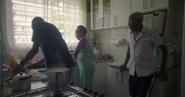 Afrika kökenli bir aile mutfakta yemek hazırlıyor, kadın ocağın yanında, yaşlı bir adam ayakta ve gözlem yapıyor, aile dinamiklerini ve günlük rutinlerini yakalıyor.
