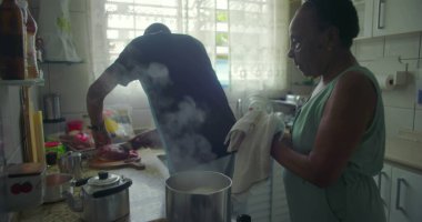 Afrika kökenli bir adam tezgahta et hazırlarken, Afrikalı bir kadın sobanın yanında iniyor. Sıradan ve samimi bir an. Günlük aile hayatını yakalıyor.
