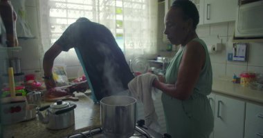 Afrika kökenli bir adam tezgahta et hazırlarken, Afrikalı bir kadın sobanın yanında iniyor. Sıradan ve samimi bir an. Günlük aile hayatını yakalıyor.