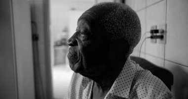 Siyah-beyaz görünümlü yaşlı bir Afro-torunu, düşünceli ve sakin bir ifadeyle yukarı bakıyor, sessiz bir ev ortamında oturuyor