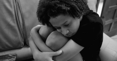 Genç Afrikalı Amerikalı çocuğun yakın çekimi, kıvırcık saçlı, derin düşüncelerle dizlerine eğilmiş, üzüntü ve iç gözlem ifade eden, kapalı alanlarda izole edilmiş, duyguları ve savunmasızlığı vurgulayan