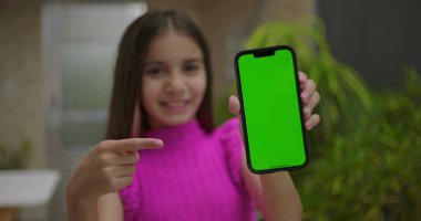 Açık hava ortamında yeşil ekran ve krom tuşlu akıllı telefona gülümseyen ve işaret eden genç kız, dijital içerik veya reklamcılık için kişiselleştirilebilir görüntüyü vurguluyor