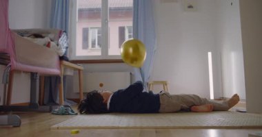 Çocuk yatak odasında yerde uzanmış yüzen altın balonla oynuyor, sakin bir konsantrasyon ve sessiz bir mutluluk gösteriyor.