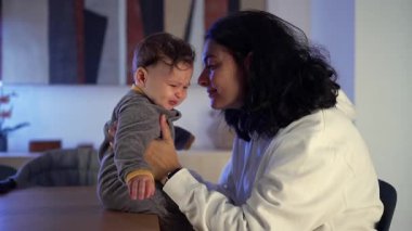 Anne evde telaşlı bir bebeği rahatlatıyor, yumuşak bir akşam ışığında küçük bir çocuğu nazikçe kucaklıyor ve onu rahatlatıyor.