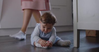 Bebeğin mutfak zemininde oturup kırıntıları merakla incelemesi, odaklanmayı ve araştırmayı göstermesi, ebeveynlerin arka planda hareket etmesi