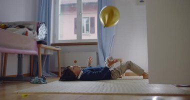 Çocuk yatak odasında yere uzanmış altın balonu havaya fırlatıyor, neşeli hayal gücü ve kaygısız neşe içinde.