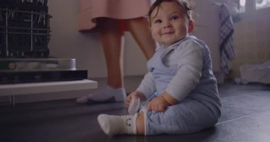 Bebeğin mutfak zemininde annesinin yanında oturması bulaşık makinesini yüklemesi, merakla gözlemlemesi ve keşfetmesi, günlük ev hayatının erken öğrenme anı