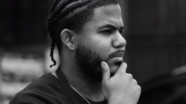 Düşünceli, saçları çenesinde örülmüş, derin düşüncelere dalmış, şehir ortamında, tek renkli yakın plan portresi olan siyah bir adamın yan profili.