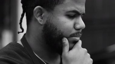 Düşünceli, saçları çenesinde örülü, derin bir yansıma, odaklanmış bir ifade, şehir dışı manzarası, tek renkli yakın plan, siyah bir adamın yan görüntüsü.