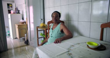 Mütevazı bir ev mutfağında, yemek masasında sessizce oturan yaşlı Afrikalı Amerikalı kadın, elini masaya koyuyor, yalnızlığı ve iç gözlem anlarını yansıtıyor.