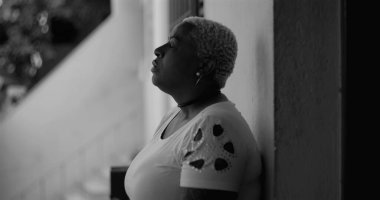 Afro-Amerikan kadın duvara yaslanmış, el kol hareketi yapıyor, umutsuzluğunu ve iç bakış açısını tek renkli bir ortamda ifade ediyor, pişmanlık ve keder temaları.