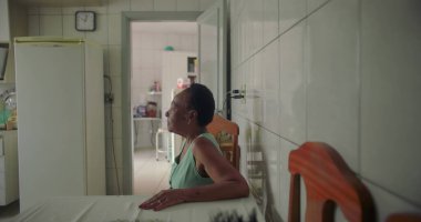 Basit bir mutfakta, yemek masasında tek başına oturan yaşlı bir kadın, düşünceli bir şekilde uzaktan bakıyor, etrafını aletler ve fayanslarla çevrili, sessiz bir anı yansıtıyor.