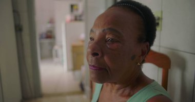 Afro-Amerikalı yaşlı bir kadın basit bir mutfakta yemek masasında oturuyor, uzaklara bakıyor, yalnızlığı ve yaşlılığın sessiz yalnızlığını yansıtıyor.