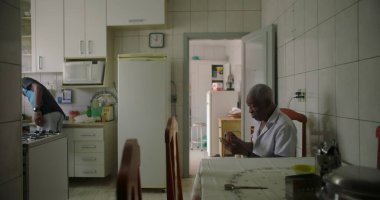 Bir yaşlı siyah adam, rahat bir mutfakta yemek masasında oturmuş, akıllı bir telefona odaklanmış, günlük rutinin ve teknolojinin özünü yakalıyor.