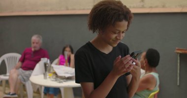 Afrika kökenli bir gencin elinde yeşil ekranlı akıllı telefon, krom anahtar özelliği gösteriliyor, arka planda çeşitli yetişkinlerin olduğu sıradan bir toplantıda gülümsüyor.