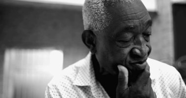 Siyah beyaz portredeki yaşlı Afro-torun adam, düşünceli ve biraz endişeli bir ifadeyle gözleri kapalı, elleri yansıtıcı bir ev ortamında çene üzerinde dinlenen