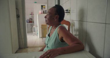 Basit bir mutfakta, yemek masasında tek başına oturan yaşlı bir kadın, düşünceli bir şekilde uzaktan bakıyor, etrafını aletler ve fayanslarla çevrili, sessiz bir anı yansıtıyor.