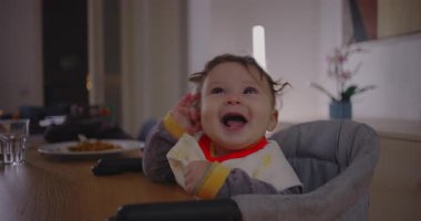 Bebek yüksek sandalyede gülüyor aile yemeği sırasında keyifle yukarı bakıyor, neşeli bebek ifadesi saf mutluluğu ve duygusal bağı yakalıyor.