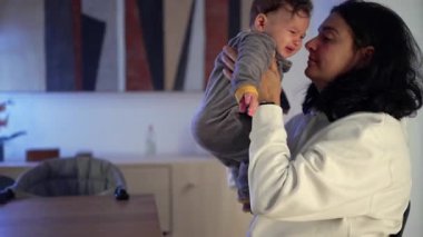 Anne bebeği masadan kaldırıyor, sıkıntı anında huzur ve rahatlığa yakın duruyor, sıcak bir iç mekanda şefkat ve duygusal bağ gösteriyor.