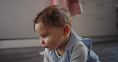 Bebeğin mutfak zemininde annesinin yanında oturması arka planda duruyor, erken öğrenme ve aile hayatı süresince merakla çevreyi keşfediyor.