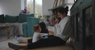 Anne, sıcak oturma odasında gün ışığıyla erken oyun zamanı etkileşimine girerken bebeğe sıcak bir şekilde gülümseyerek yerde oturuyor.