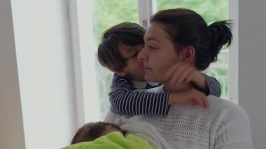Anne, çocukları pencerenin yanında kucaklıyor, büyük oğul bebeği tutarken yanağını öpüyor, anne sevgisinin ve aile sıcaklığının şefkatli ifadesi.