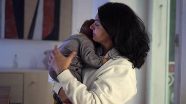 Anne bebeği göğsüne yakın tutuyor, çocuğu rahatlatıyor ve sallıyor. Ağladıktan sonra sakinleşiyor. Sıcak bir akşamın ışığı altında şefkatli bir an.