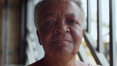 Tarafsız bir ifadeyle doğrudan kameraya bakan yaşlı bir kadının portresi, evdeki sakinlik anında doğal ışık altında demir parmaklıkların yanında duruyor.