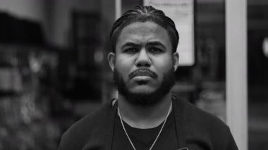 Saçları örgülü ciddi bir siyah adamın portresi doğrudan kameraya bakıyor, kentsel arka plan, yoğun bakış, güçlü ifade, düşünceli tavır, tek renkli yakın çekim