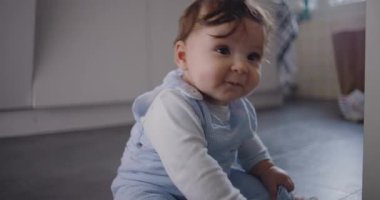 Bebek mutfak zemininde oturup komik suratlar yapıyor. Erken öğrenme ve sıcak aile ortamında keşifler sırasında sevimli bir merak ve eğlence ifadesi sergiliyor.