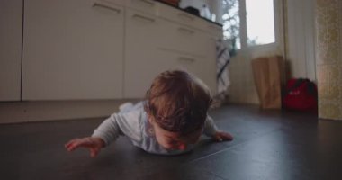 Bebek, erken motor beceri denemesi sırasında hayal kırıklığı içinde mutfakta yatar, anne tarafından alınmadan önce duygularını ve çabalarını ifade eder.